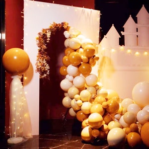 May include: A white backdrop with a balloon arch made of white, cream, and gold balloons. The arch is decorated with dried flowers and greenery. There is a large gold balloon on the left side of the image.