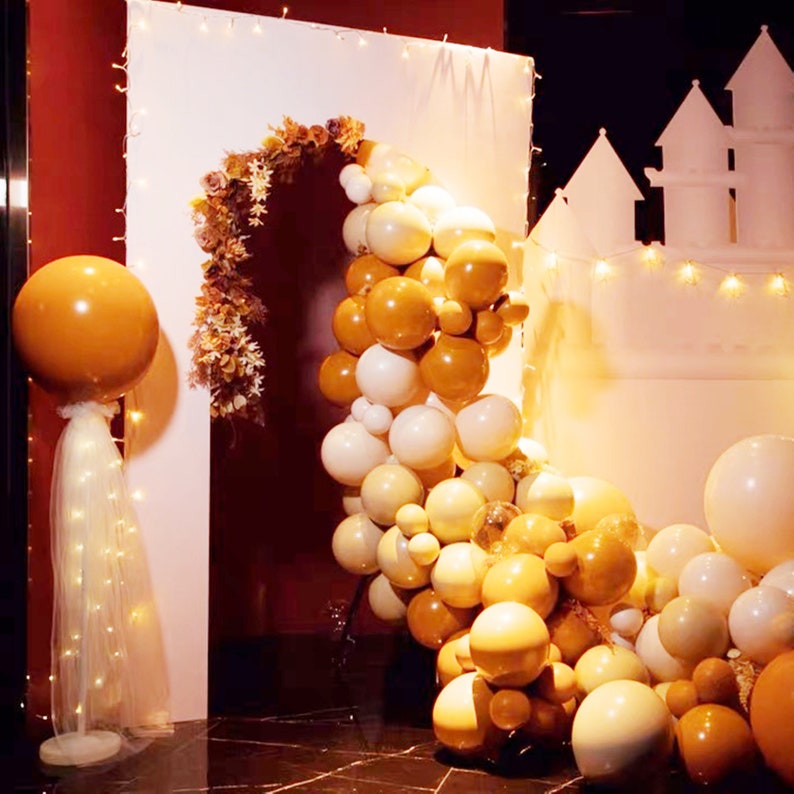 May include: A white backdrop with a balloon arch made of white, cream, and gold balloons. The arch is decorated with dried flowers and greenery. There is a large gold balloon on the left side of the image.
