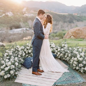 May include: A couple embraces outdoors, framed by white flowers and a mountain backdrop. The woman wears a long, white lace wedding dress. The man is in a dark suit. They stand on a rug with a blue pillow, with the sun setting in the background.