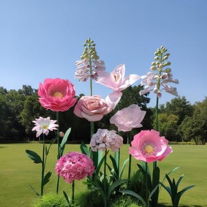 May include: A vibrant display of large, paper flowers in shades of pink, white, and green. The arrangement includes roses, poppies, and other blooms, set against a backdrop of green grass and a clear blue sky. The flowers are arranged in a garden setting.