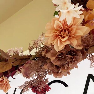 May include: A floral garland with pink, brown, and white flowers and leaves. The garland is hanging from a ceiling and is in front of a white sign with black text.