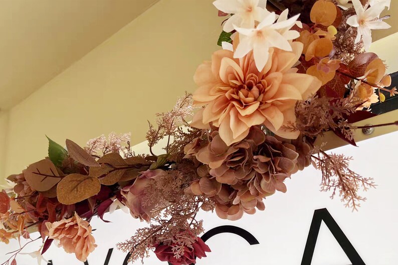 May include: A floral garland with pink, brown, and white flowers and leaves. The garland is hanging from a ceiling and is in front of a white sign with black text.