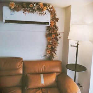 May include: A brown leather sofa with a white pillow in front of a white wall. A garland of dried flowers hangs on the wall above the sofa. A black floor lamp with a white shade is in the corner of the room.