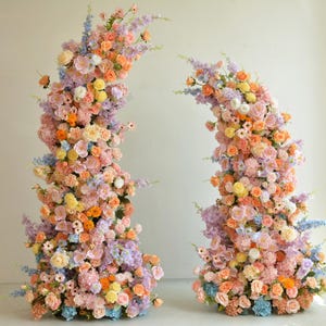 Puede incluir: Dos grandes arreglos florales en forma de media luna. Los arreglos están hechos de una variedad de flores pastel, incluyendo rosa, morado, amarillo y naranja.