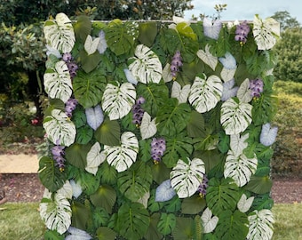 Puede incluir: Un exuberante muro verde artificial con una variedad de hojas tropicales, incluyendo grandes monstera de hojas divididas y hojas más pequeñas en forma de corazón. Acentos de flores blancas y lavanda se intercalan en el follaje.