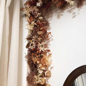 May include: A brown and cream floral garland with white flowers and leaves, hanging on a white wall next to a brown wooden mirror.