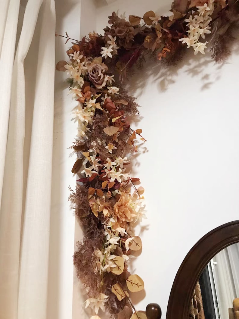 May include: A brown and cream floral garland with white flowers and leaves, hanging on a white wall next to a brown wooden mirror.