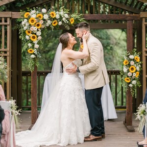 May include: A wedding ceremony featuring a person in a white strapless lace gown with a long veil and a person in a tan blazer and dark trousers standing under a wooden arbor decorated with sunflowers and white roses, facing each other.