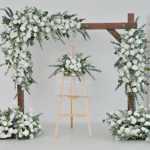 May include: A wooden archway decorated with white flowers and greenery. The archway is set up for a wedding ceremony or other special event. A sign on an easel reads "welcome to our day".