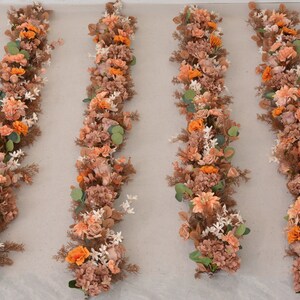 May include: Four long garlands of artificial flowers in shades of orange, brown, and white. The garlands are arranged vertically on a light beige background.