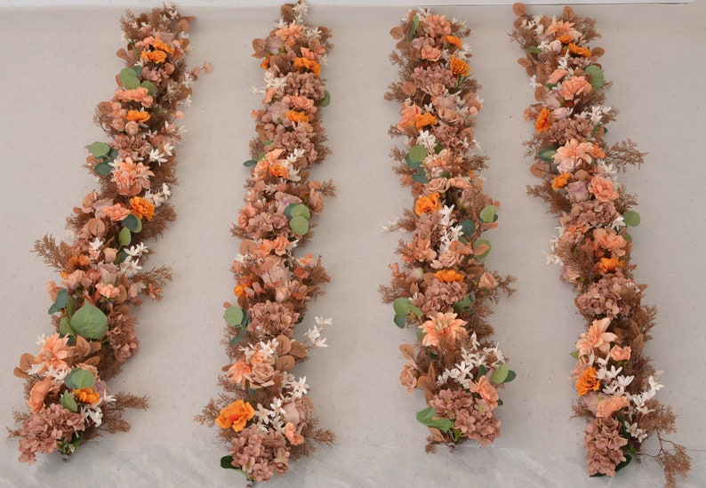 May include: Four long garlands of artificial flowers in shades of orange, brown, and white. The garlands are arranged vertically on a light beige background.