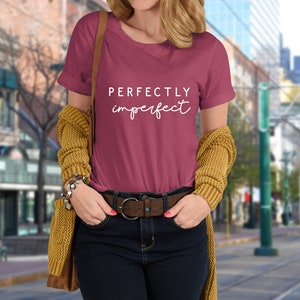 May include: A maroon t-shirt with the text "PERFECTLY imperfect" in white lettering. The shirt is being worn by a person in a city setting.