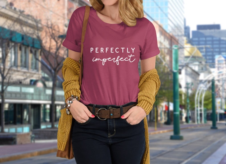 May include: A maroon t-shirt with the text "PERFECTLY imperfect" in white lettering. The shirt is being worn by a person in a city setting.