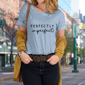 May include: A light blue t-shirt with the text "PERFECTLY imperfect" in black lettering. The shirt is being worn by a person with a brown leather belt and a yellow cardigan.