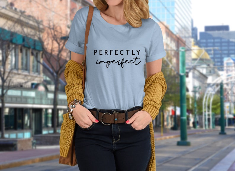 May include: A light blue t-shirt with the text "PERFECTLY imperfect" in black lettering. The shirt is being worn by a person with a brown leather belt and a yellow cardigan.