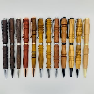 Puede incluir: Una colección de doce bolígrafos de madera hechos a mano en varios tonos de marrón y beige. Cada bolígrafo presenta un diseño único, con detalles metálicos plateados, cobrizos y dorados. Los bolígrafos están dispuestos en fila sobre un fondo blanco.