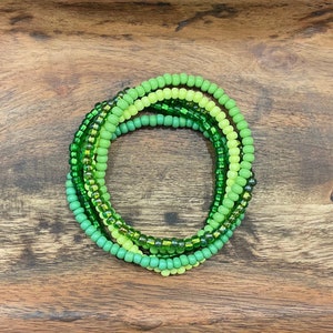 May include: A set of five green and lime green beaded bracelets stacked on top of each other. The bracelets are made of small, round beads.