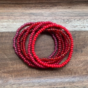 May include: A set of red beaded bracelets in various shades of red, stacked on top of each other on a wooden surface.