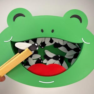 May include: A green frog-shaped container with a wide open mouth revealing a red tongue and multiple black and white fly-shaped pieces inside. A pair of light brown wooden tweezers holds one black and white fly-shaped piece near the frog's mouth.