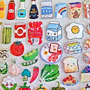 May include: A collection of colorful, cartoon-style food stickers, including a carton of milk, a fried egg, a slice of toast with bacon, and various fruits and vegetables. The stickers have a glittery effect.