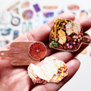 May include: Three holographic stickers featuring a cutting board with a slice of orange, a loaf of bread, and a charcuterie board with cheese, grapes, and crackers.