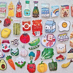 May include: A collection of colorful, cartoon-style food and drink stickers, including a banana, watermelon slice, milk carton, egg, and popcorn. The stickers have a glittery effect and smiling faces.