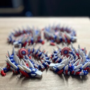 May include: Four articulated dragon toys in red, white, and blue, with star and stripe patterns. The toys have white spikes and are arranged on a wooden surface. The design evokes a patriotic theme.