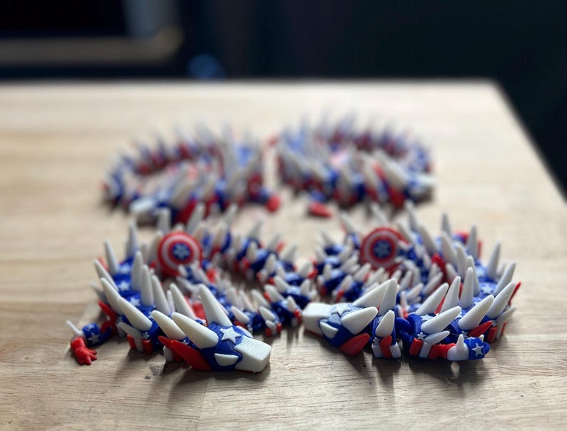 May include: Four articulated dragon toys in red, white, and blue, with star and stripe patterns. The toys have white spikes and are arranged on a wooden surface. The design evokes a patriotic theme.