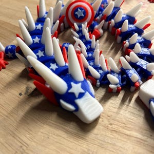 May include: A 3D-printed articulated dragon toy in red, white, and blue. The dragon features a patriotic design with stars and stripes, and white spikes along its back. A small shield with a star is also visible.