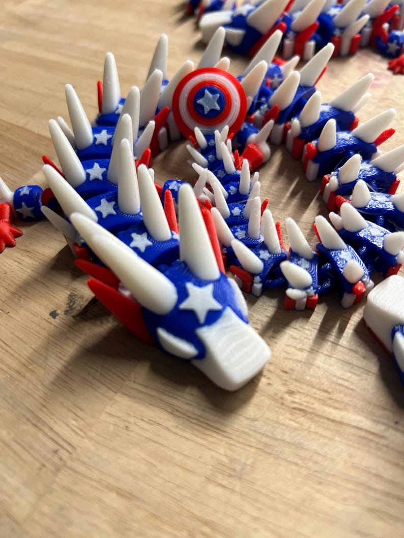 May include: A 3D-printed articulated dragon toy in red, white, and blue. The dragon features a patriotic design with stars and stripes, and white spikes along its back. A small shield with a star is also visible.