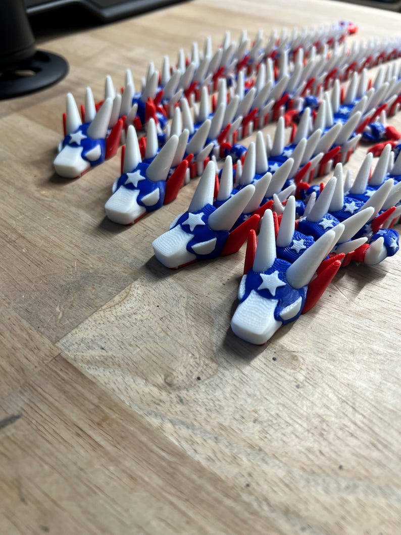 May include: A collection of red, white, and blue dragon-shaped ornaments. The dragons have white spikes, red wings, and blue heads with white stars. The ornaments are arranged on a wooden surface.