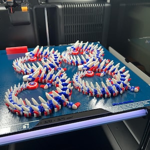 May include: Several 3D-printed articulated dragon toys in red, white, and blue, resembling the American flag. The toys are arranged on a blue surface, with a 3D printer in the background. The toys are made of plastic and have a spiky design.