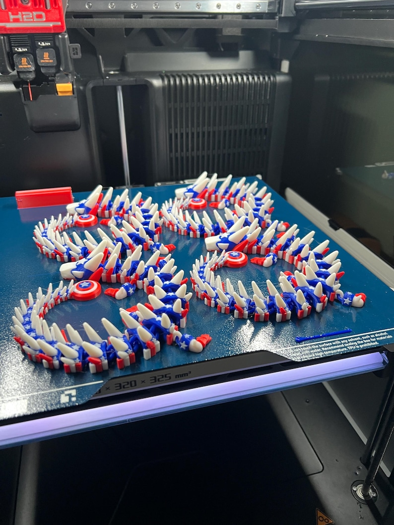 May include: Several 3D-printed articulated dragon toys in red, white, and blue, resembling the American flag. The toys are arranged on a blue surface, with a 3D printer in the background. The toys are made of plastic and have a spiky design.