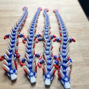 May include: Several articulated dragon toys in red, white, and blue. The dragons have a blue body with white star accents, white spikes, and red claws and accents. The toys are arranged on a wooden surface.
