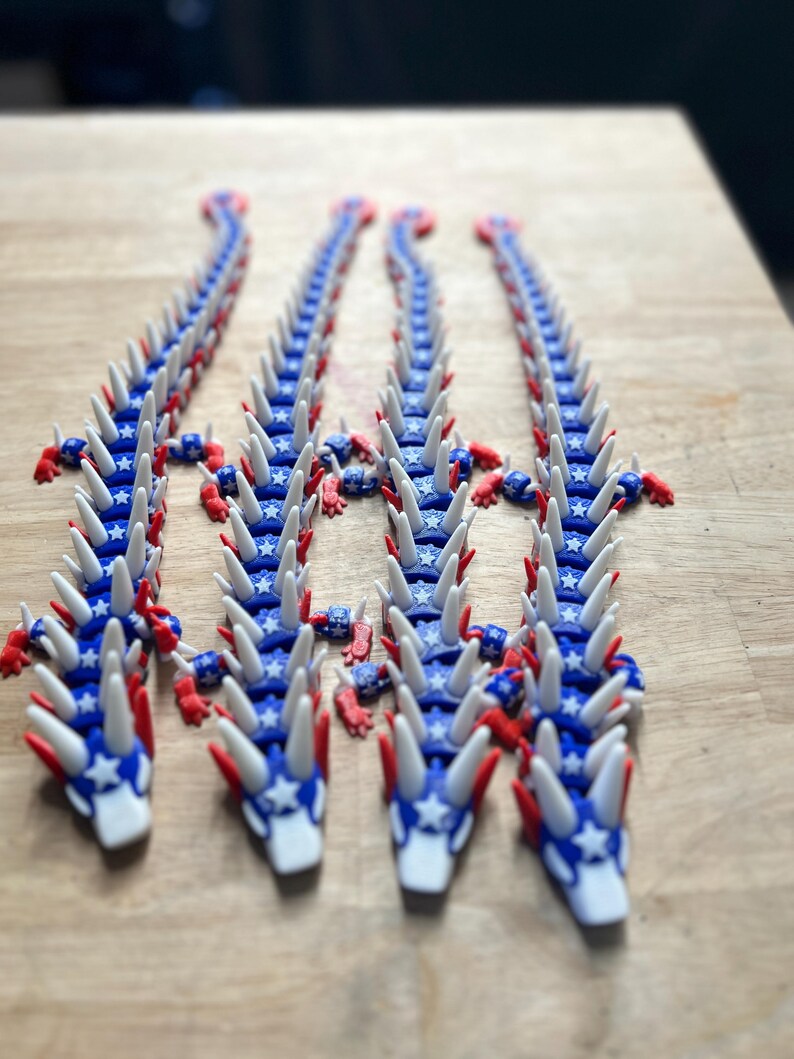 May include: Several articulated dragon toys in red, white, and blue. The dragons have a blue body with white star accents, white spikes, and red claws and accents. The toys are arranged on a wooden surface.
