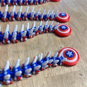 May include: Several 3D-printed articulated toys in red, white, and blue, resembling a segmented creature. Each segment has a blue and white design with red accents. The toys end with a circular shield design with a star in the center.