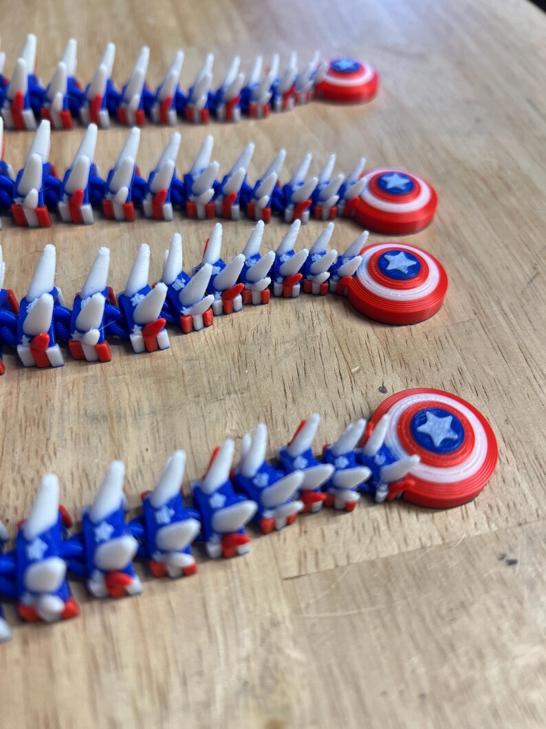 May include: Several 3D-printed articulated toys in red, white, and blue, resembling a segmented creature. Each segment has a blue and white design with red accents. The toys end with a circular shield design with a star in the center.