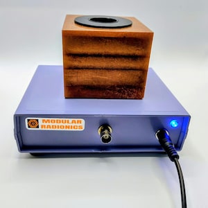 May include: A light purple electronic device labeled Modular Radionics with a BNC connector, a power cable, and a glowing blue light. A brown, textured, cube-shaped block with a circular black insert sits on top of the rectangular purple unit.