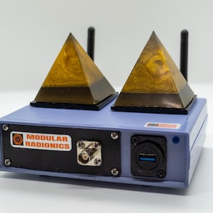 May include: A light blue electronic device with two golden pyramid-shaped structures on top. The device has a black panel with the text "MODULAR RADIONICS" and various ports and connectors. Two black antennas are also present.