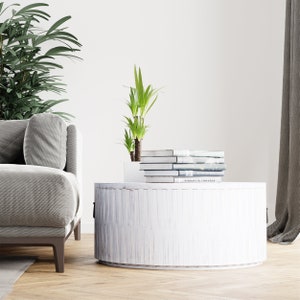 May include: White round wooden coffee table with a stack of books on top. The table has a distressed white finish and a slightly rounded edge. A green plant is in a white planter behind the books.