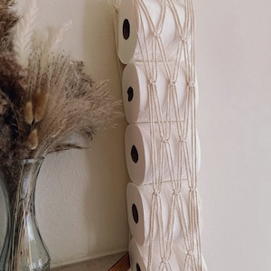 May include: A white macrame wall hanging with a wooden dowel at the top. The macrame is designed to hold toilet paper rolls, with five rolls hanging in the net. The macrame has a fringe at the bottom.
