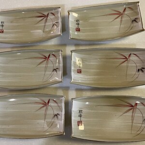 May include: Set of six rectangular ceramic plates. Each plate features a cream-colored background with a bamboo design in red and black. The plates have a gold trim and Japanese characters.