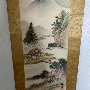 May include: A traditional Japanese scroll painting depicting a serene landscape. The artwork features a snow-capped mountain, a river, and a small house with a bridge. The scroll is framed with a gold patterned border.
