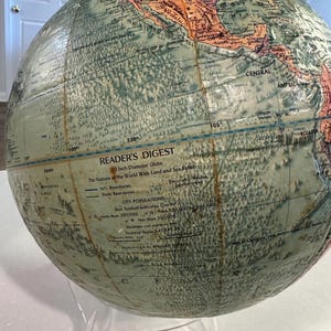 May include: A vintage Reader's Digest 12-inch diameter globe with land and sea relief. The globe features a green and brown color scheme with a detailed map of the world. The globe is on a clear acrylic stand.