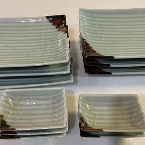 May include: A collection of square and rectangular ceramic plates. The plates are light green with a textured, ribbed design and a dark brown accent on one corner. The set includes both large and small plates.