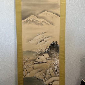 May include: A traditional Japanese scroll painting depicting a snowy mountain landscape with a small village in the foreground. The painting is done in shades of gray and brown, with hints of white for the snow. The scroll is mounted on a gold background with a decorative border.