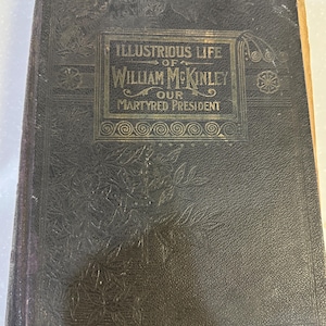 May include: An antique book with a dark cover, embossed with floral patterns. The title, "Illustrious Life of William McKinley, Our Martyred President," is displayed in gold lettering within a decorative frame. The book's edges show signs of age.