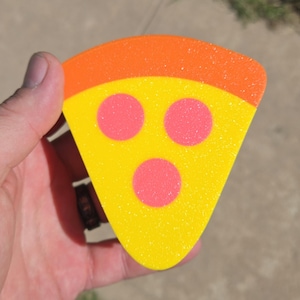 Puede incluir: Un adorno en forma de porción de pizza triangular con un borde naranja texturizado en la parte superior, una sección de queso amarillo brillante y tres ingredientes circulares rosas dispuestos en patrón triangular.