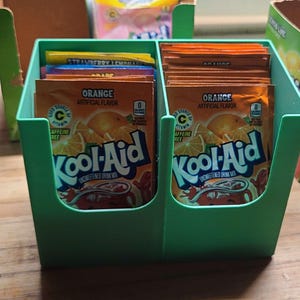 Puede incluir: Un dispensador de plástico verde con dos compartimentos, cada uno con una pila de sobres de mezcla para bebidas Kool-Aid de color naranja. Los sobres tienen el texto "Sabor artificial a naranja" y "Kool-Aid Mezcla de bebida no endulzada".