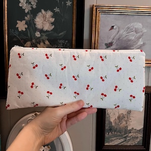 May include: A white zippered pouch with a red cherry print on a white background. The pouch is held in a hand.